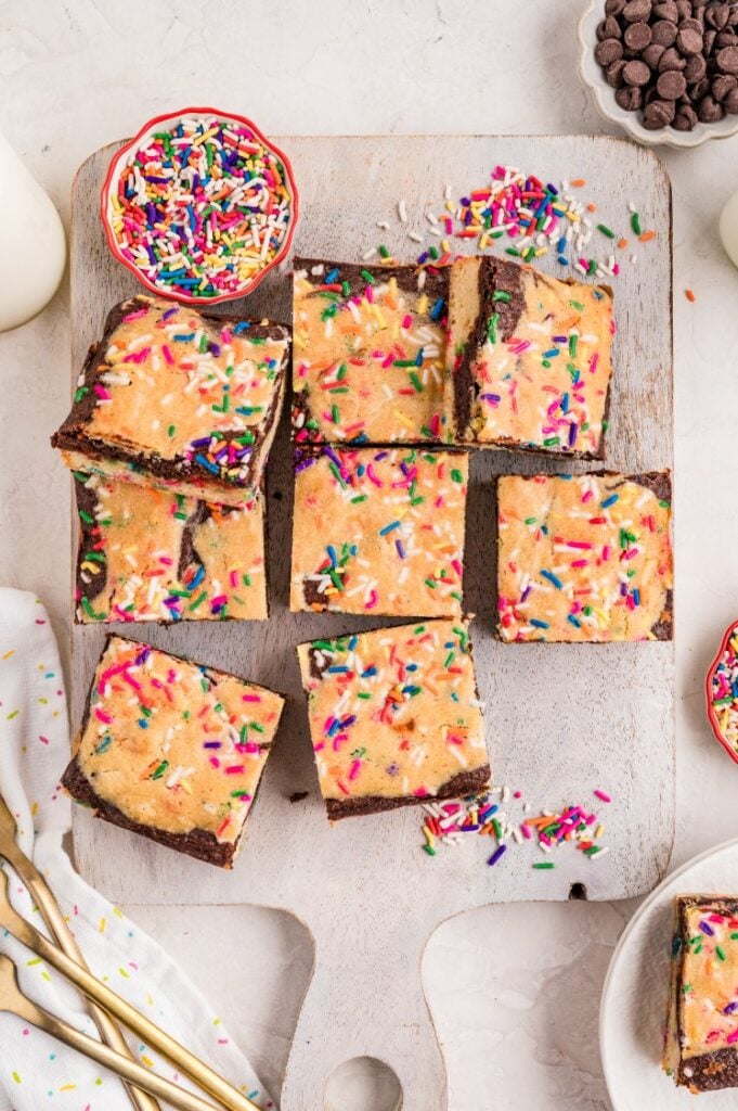 Overhead view of sliced birthday brownies on a wooden board, each topped with colorful rainbow sprinkles. A small bowl of extra sprinkles sits nearby along with chocolate chips, gold forks, and a white cloth with scattered sprinkles.