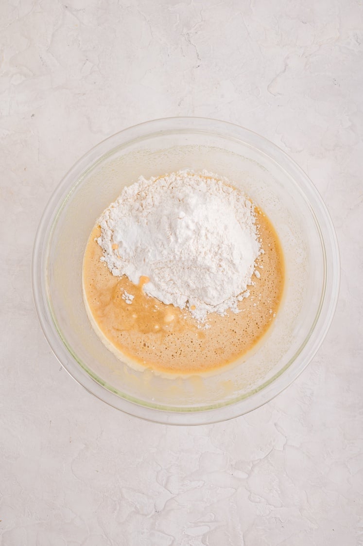 A glass mixing bowl filled with wet brownie ingredients, topped with a mound of all-purpose flour before being mixed together.