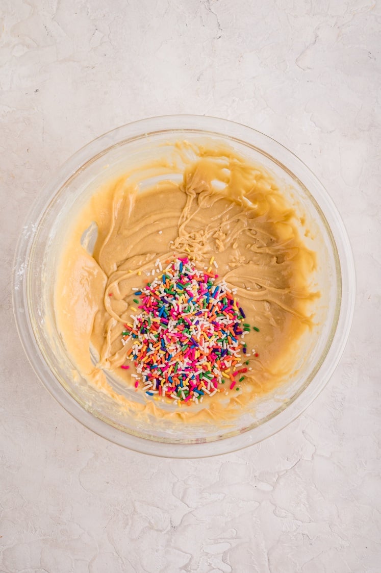 A glass mixing bowl filled with thick vanilla cake batter, topped with a pile of colorful rainbow sprinkles ready to be folded in.