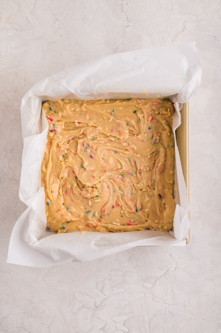 Square baking pan lined with parchment paper and filled with colorful funfetti cake batter for birthday brownies before baking.