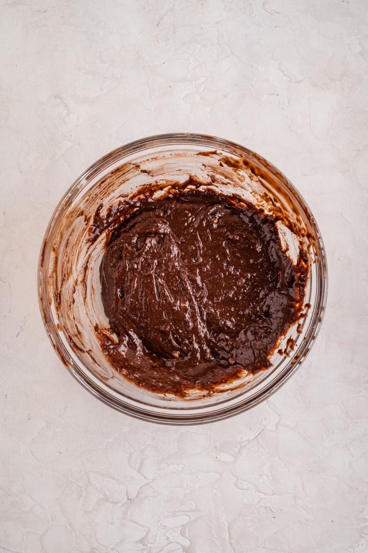 Glass bowl filled with thick, freshly mixed chocolate brownie batter on a light textured surface.