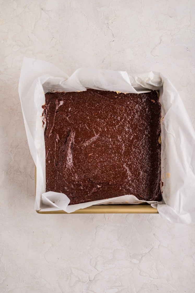 Chocolate brownie batter spread evenly over the blondie layer in a parchment-lined baking pan.