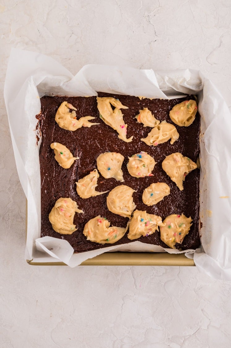 Pan of brownie batter topped with scattered dollops of funfetti cookie dough, ready to be swirled and baked.