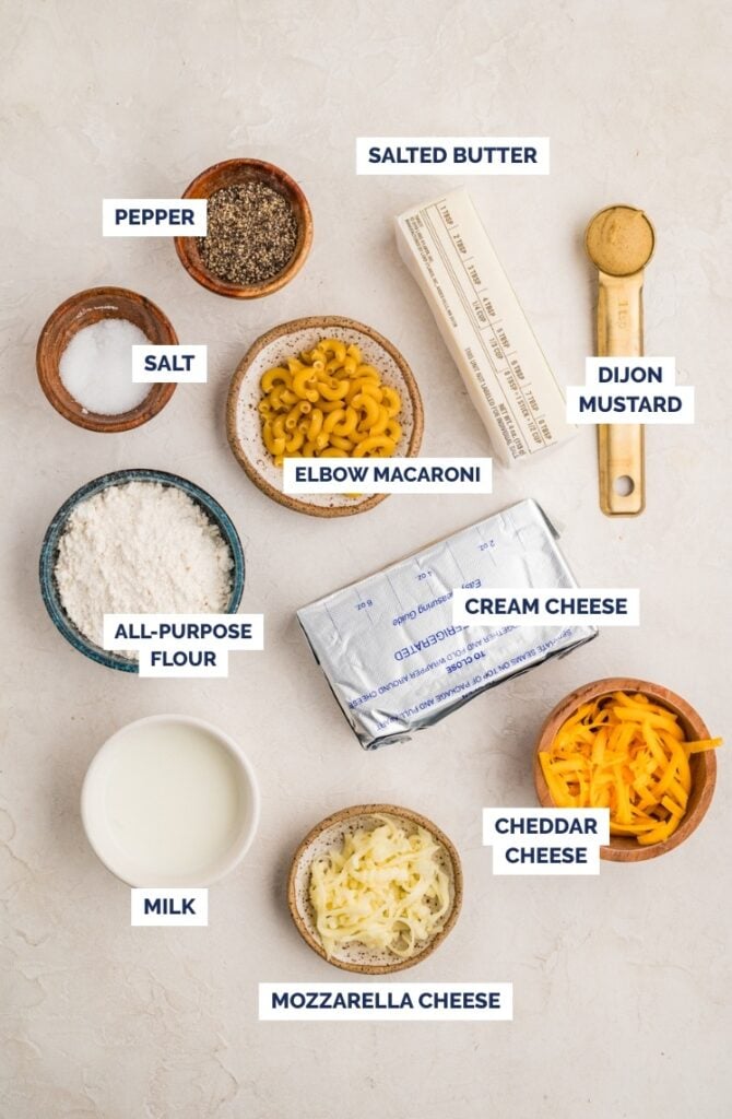 Ingredients for macaroni and cheese arranged on a light surface: pepper, salt, all-purpose flour, milk, elbow macaroni, salted butter, Dijon mustard, cream cheese, cheddar cheese, and mozzarella cheese.