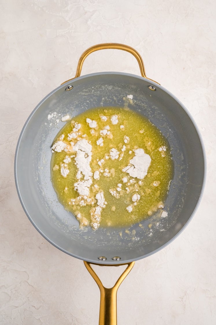 A gray and gold pan on a light surface contains melted butter with clumps of flour being mixed in.