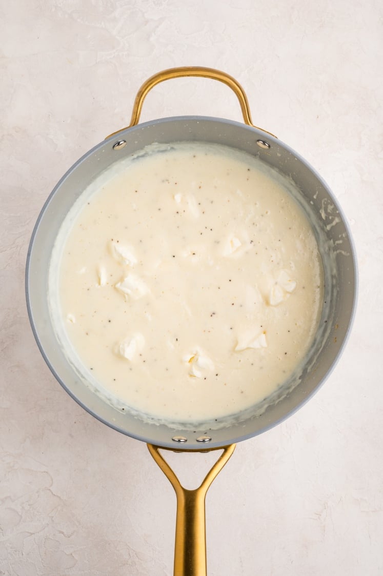 A saucepan with a golden handle contains creamy white sauce with visible chunks, set against a light, textured background.