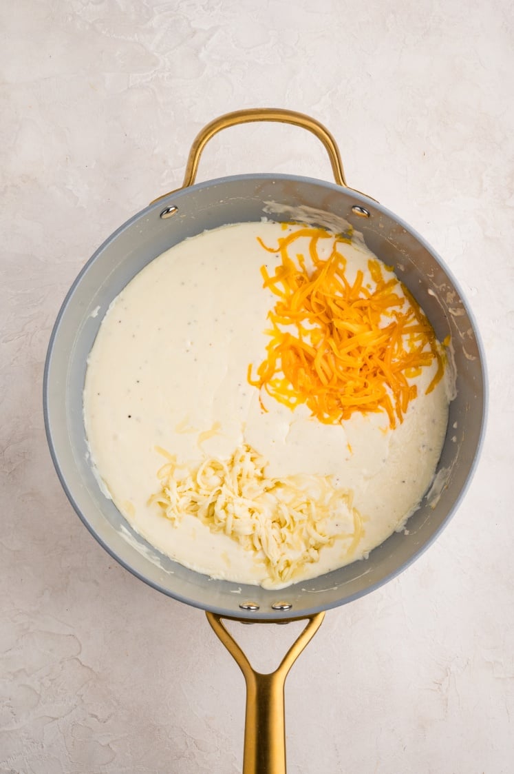 A gold-handled saucepan contains creamy white sauce with shredded yellow and white cheese on top, ready to be mixed in. The pan sits on a light, textured surface.