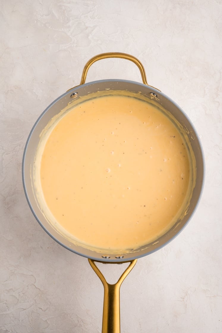 A saucepan with a gold handle contains creamy, light orange cheese sauce, viewed from above on a light textured surface.