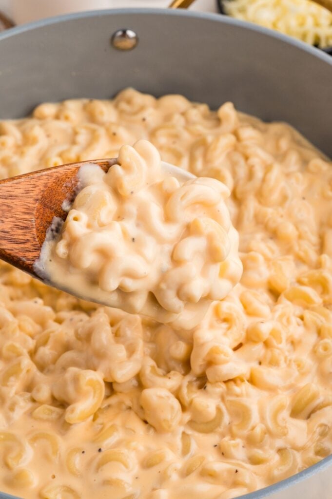 A wooden spoon scoops creamy macaroni and cheese from a pot filled with elbow macaroni coated in a rich, melted cheese sauce.