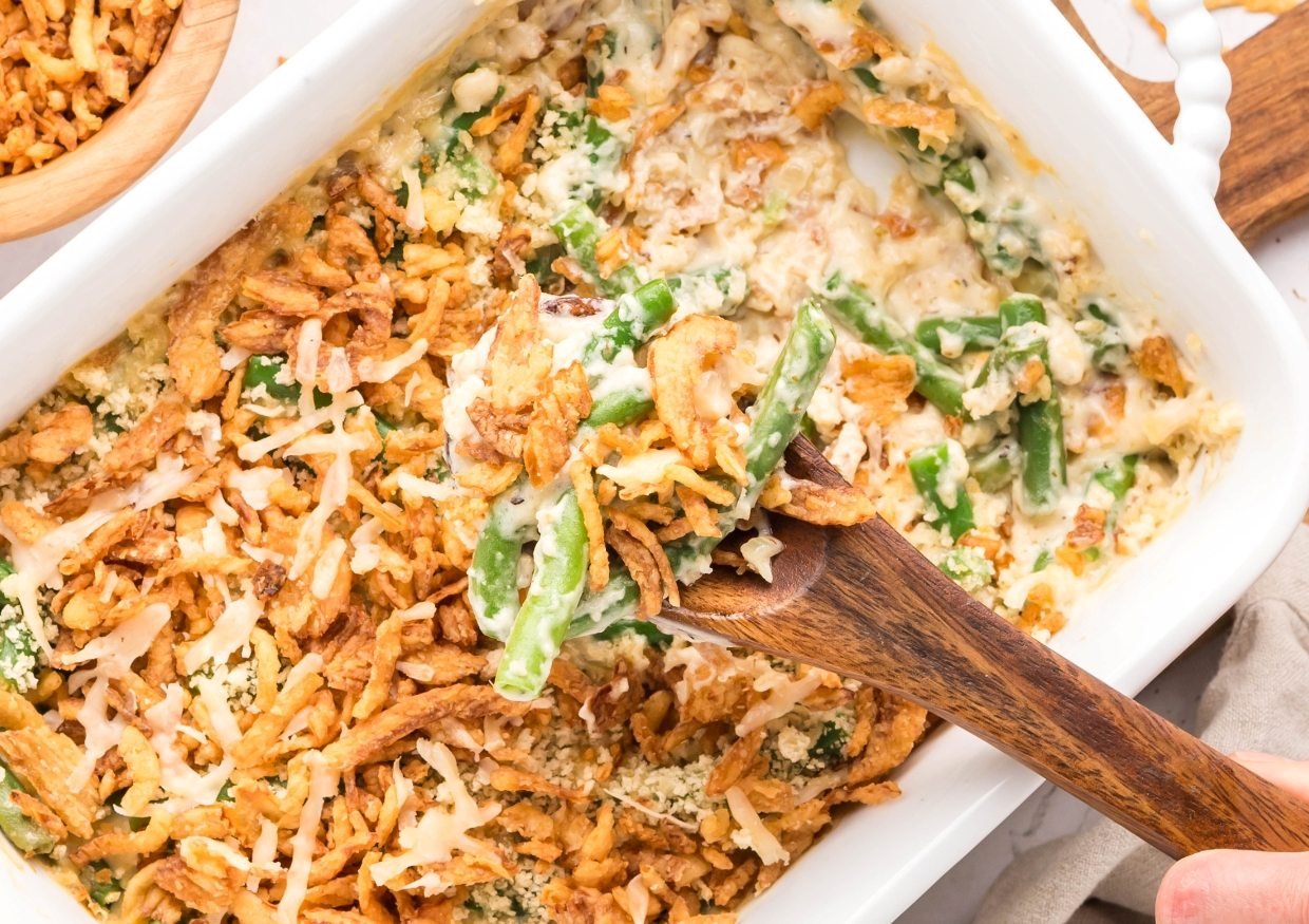 A serving of green bean casserole with crispy fried onions being scooped from a white baking dish with a wooden spoon.