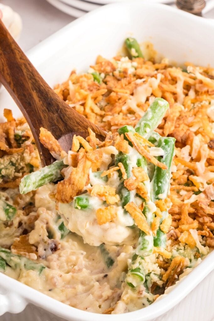 A wooden spoon scooping creamy fresh green bean casserole topped with crispy fried onions from a white baking dish.