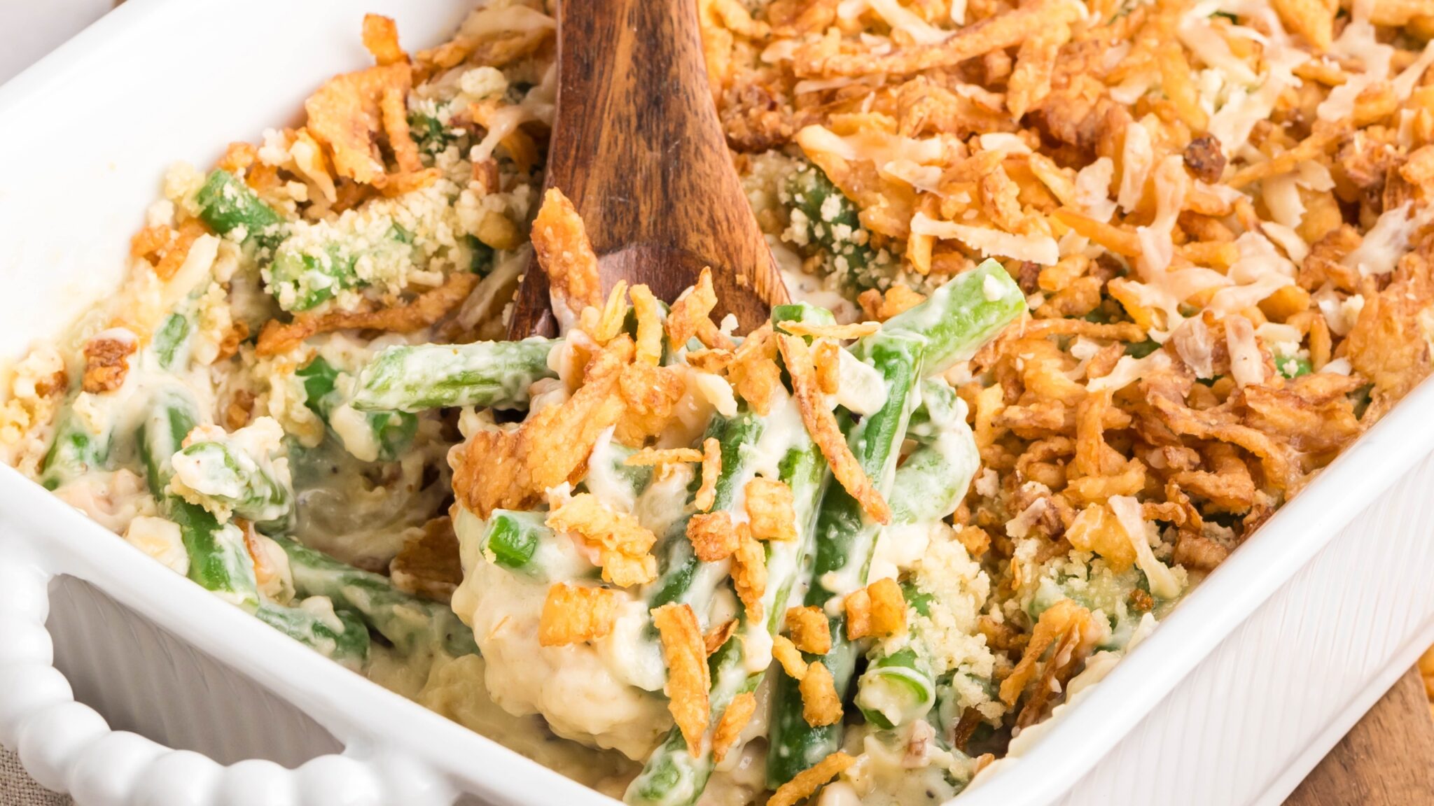 Partially scooped green bean casserole in a white ceramic baking dish with a wooden spoon, showing crispy onion topping and creamy interior.