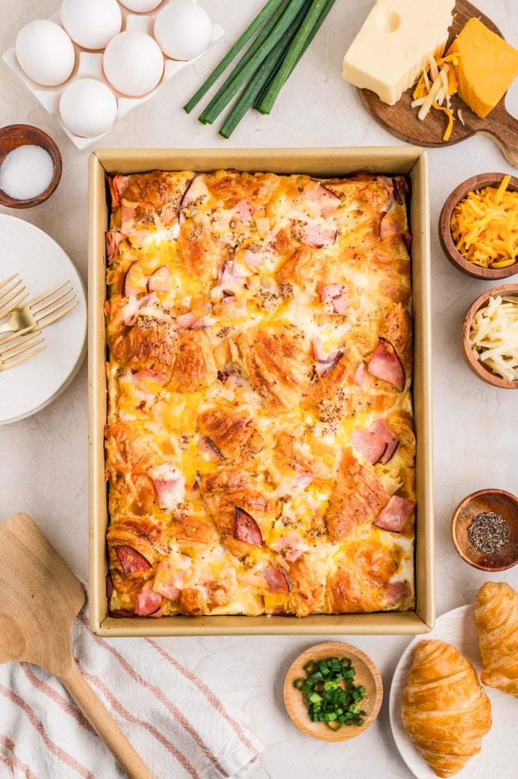 Golden-brown ham and cheese croissant breakfast bake in a pan, surrounded by ingredients like eggs, cheese, and green onions.