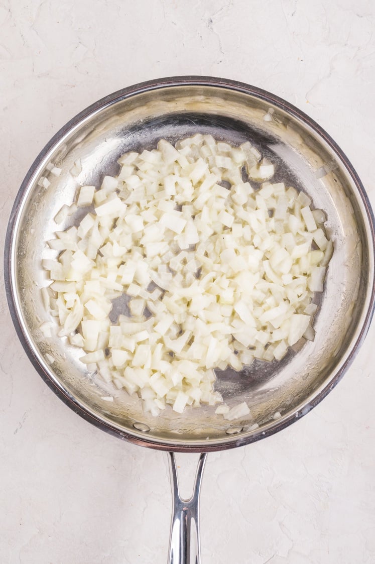 Chopped onions being sautéed in a stainless steel pan on a light-colored surface, perfectly prepped for adding to your savory Ham and Cheese Croissant Bake.