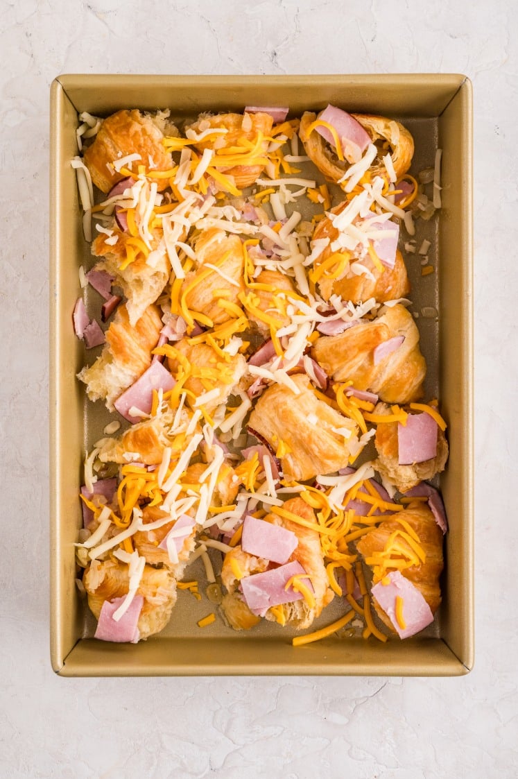 Torn croissant pieces layered in a baking dish and topped with shredded cheddar, shredded Swiss cheese, and chunks of black forest ham, ready to be baked for a ham and cheese croissant breakfast casserole.
