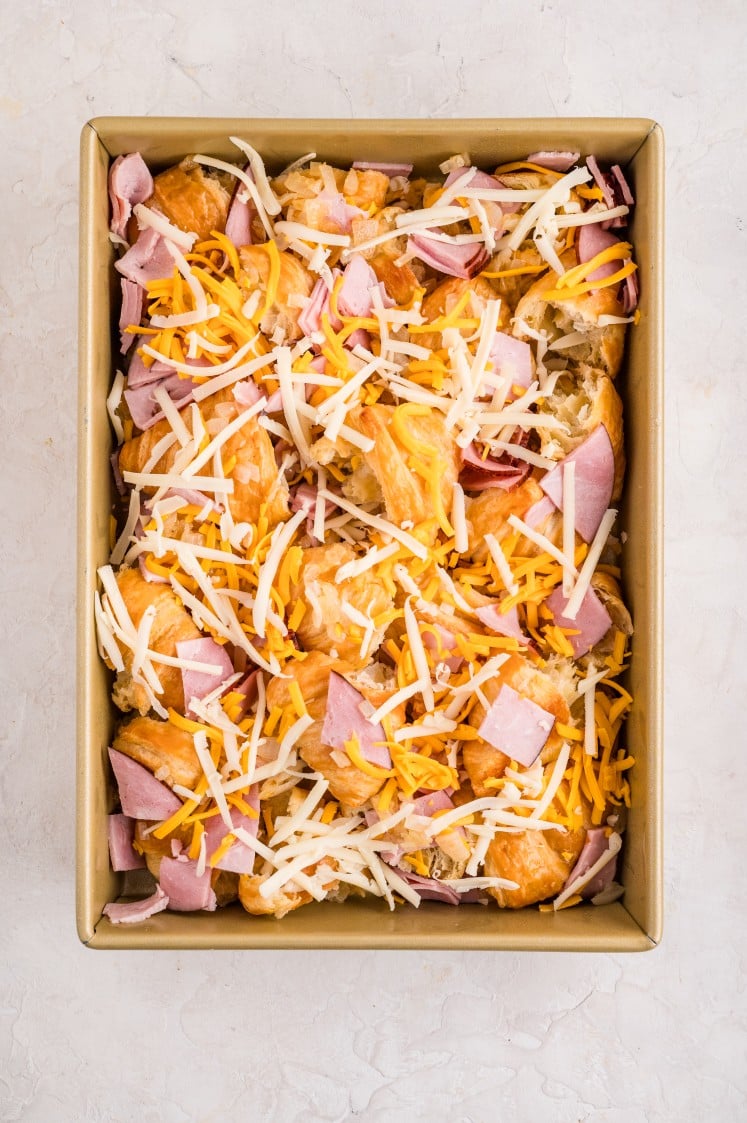 Top-down view of a baking pan filled with torn croissant pieces layered with chopped ham and shredded cheddar and Swiss cheese, ready to be baked for a ham and cheese croissant breakfast casserole.