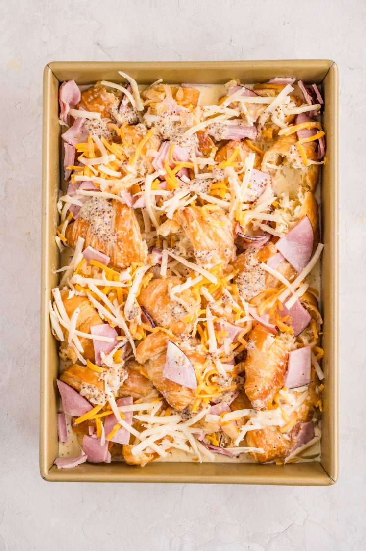 Unbaked Ham and Cheese Croissant Breakfast Bake in a rectangular pan, filled with torn croissants layered with chopped ham, shredded cheddar and Swiss cheese, and whole-grain mustard before adding the egg mixture.