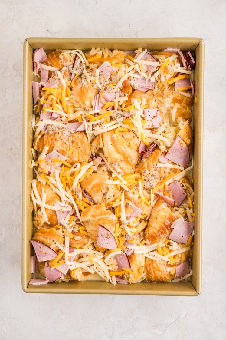 A rectangular baking dish filled with an unbaked Ham and Cheese Croissant Bake made of croissant pieces, shredded cheese, and chopped ham.