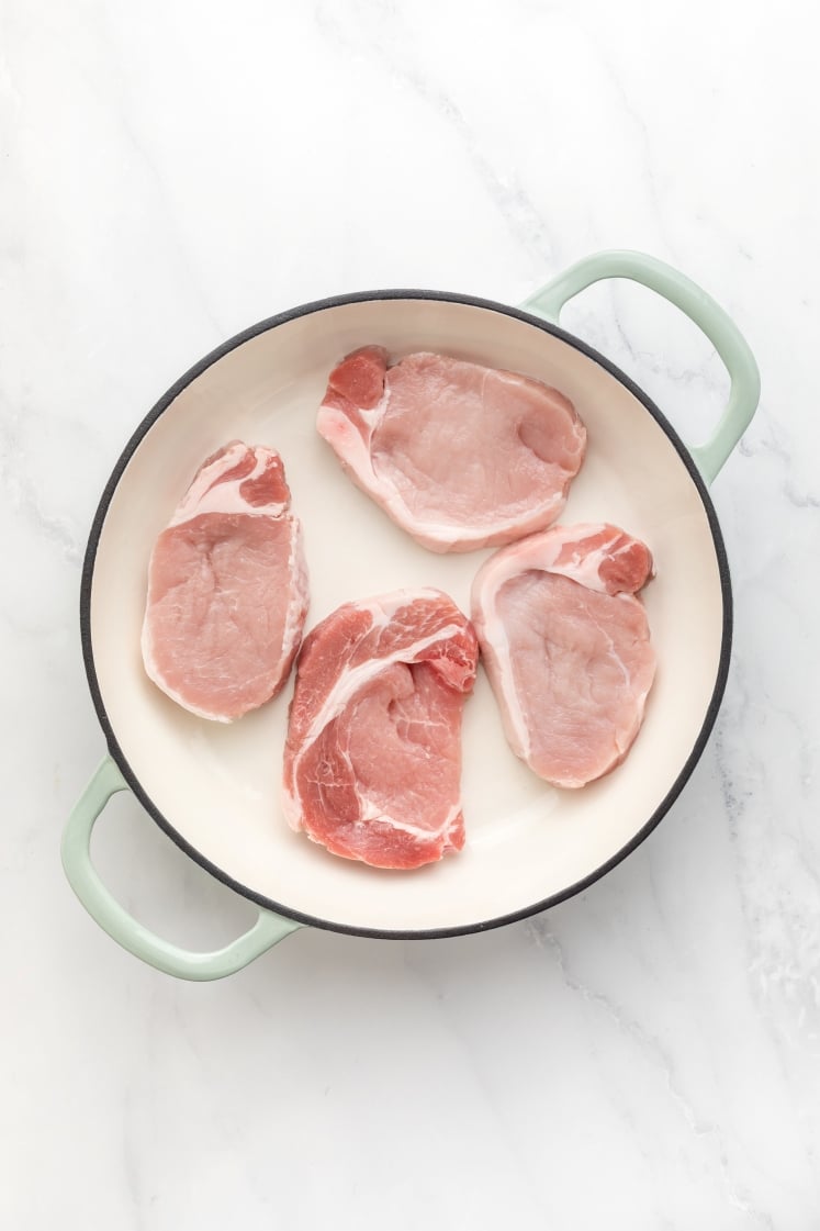 Four raw bone-in pork chops arranged in a large white skillet with green handles, ready to be cooked.
