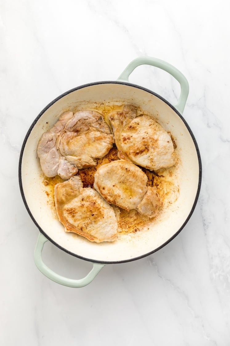 Seared pork chops in a large white skillet with browned edges and pan drippings, showing the meat after the initial browning step.