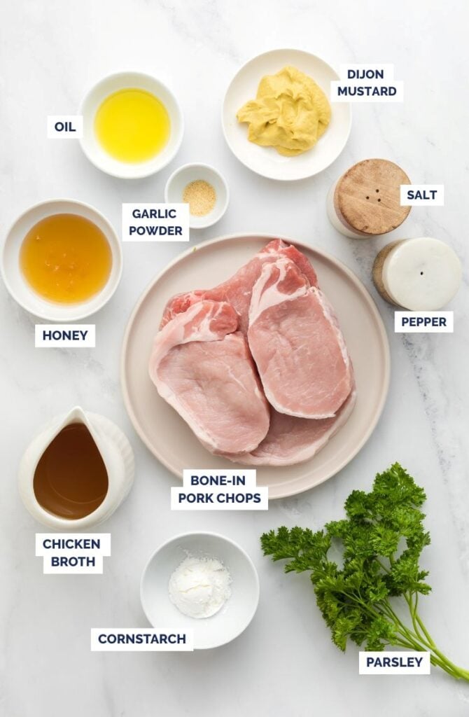 A flat lay of ingredients for honey mustard pork chops, including bone-in pork chops, Dijon mustard, honey, chicken broth, oil, garlic powder, cornstarch, fresh parsley, and salt and pepper shakers, each labeled clearly.