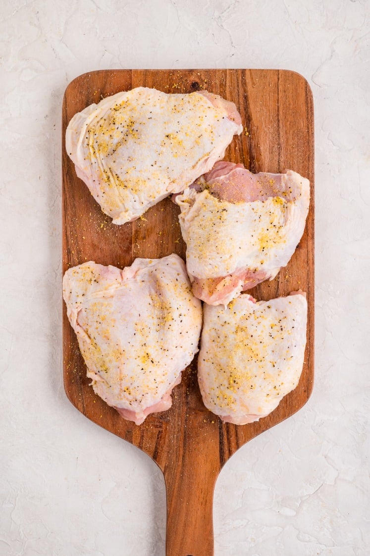 Raw chicken thighs seasoned with lemon pepper and salt on a wooden cutting board, ready to be cooked for lemon chicken rice.