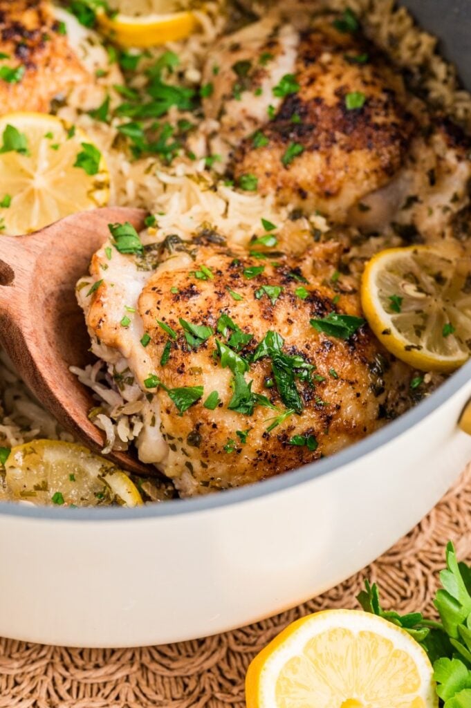 Close-up of lemon chicken rice featuring browned chicken thighs, fresh parsley, and sliced lemons with a wooden spoon serving a portion.