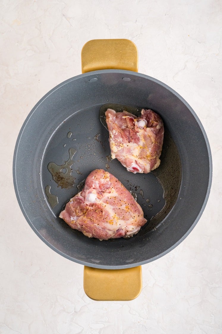 Raw seasoned chicken thighs searing in a gray pot with oil, starting to brown for lemon chicken rice.