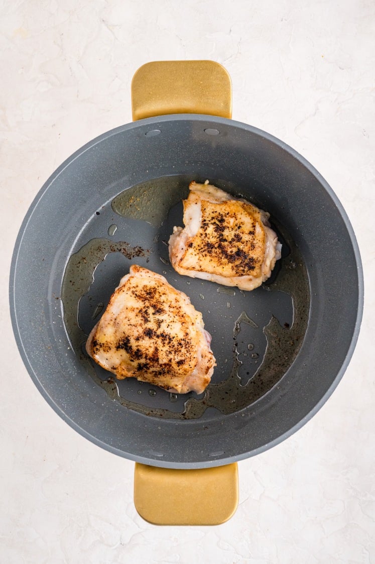 Golden-browned chicken thighs searing in a large gray pot with oil, showing crispy seasoned skin during the cooking process for lemon chicken rice.