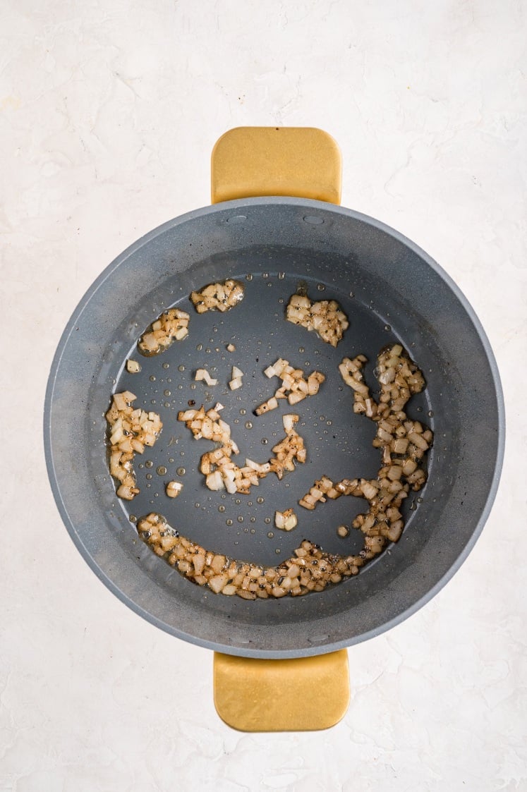 Large gray pot with sautéed diced onions cooking in oil, lightly browned and softened, sitting on a white surface with a gold handle visible.