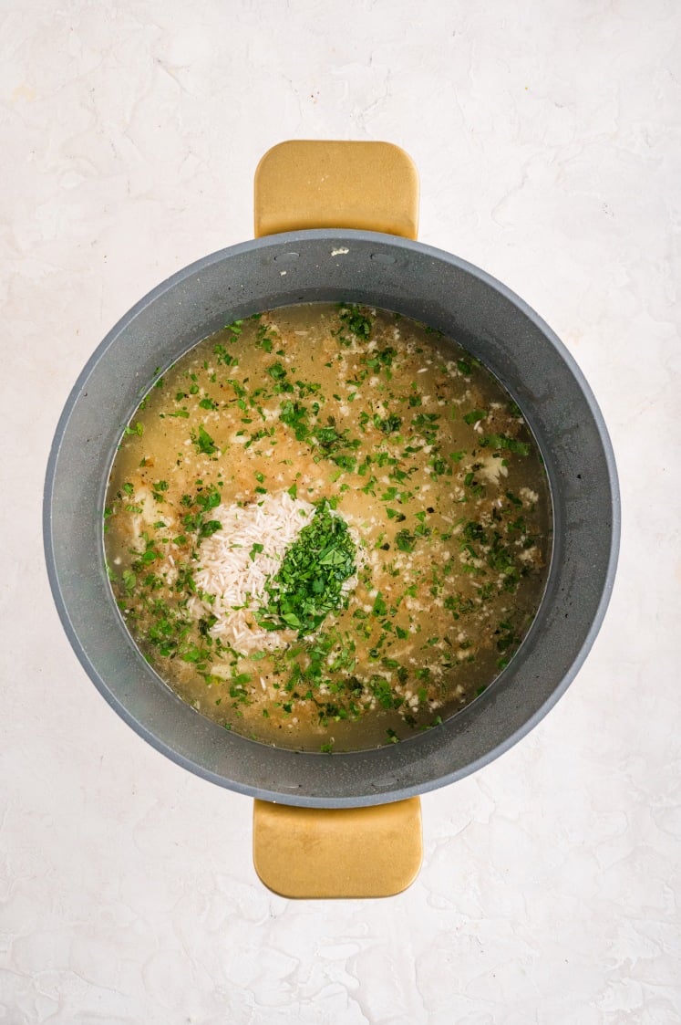 Large pot filled with broth, uncooked long-grain rice, garlic, and fresh parsley for a one-pot lemon chicken rice recipe, shown before simmering.