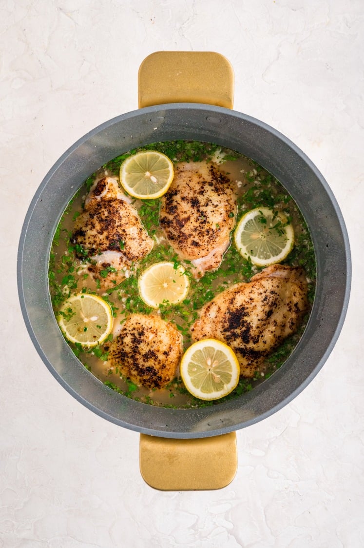 Large pot filled with seared chicken thighs simmering in broth with sliced lemons and chopped parsley, preparing lemon chicken rice on a stovetop.
