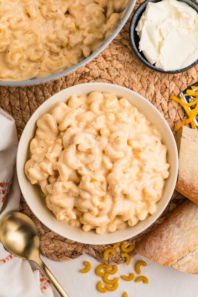 A bowl of creamy macaroni and cheese sits on a woven mat next to a spoon, a bread roll, uncooked pasta, and a small dish of cheese.
