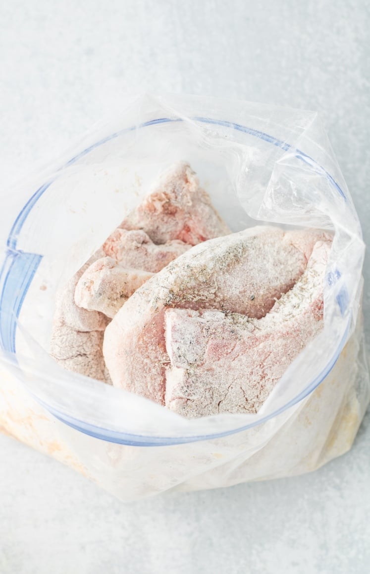 Raw pork chops coated in seasoned flour inside a zip-top plastic bag, ready to be cooked. The meat is lightly dusted and arranged on a light gray surface.