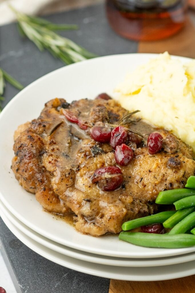 Plated maple cranberry pork chop topped with rosemary and tart cranberries, served alongside creamy mashed potatoes and fresh green beans.