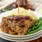Maple cranberry sauce being poured over a tender pork chop served with mashed potatoes and green beans, creating a rich and savory holiday-style meal.