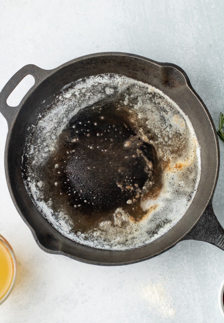 Overhead view of a cast-iron skillet with melted butter starting to brown and bubble, preparing to cook on a light gray surface.