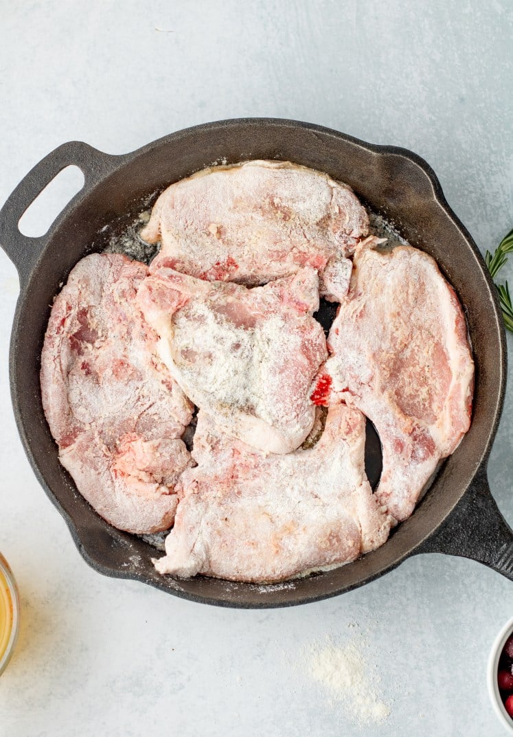 Floured raw pork chops arranged in a single layer in a cast-iron skillet, ready to be seared. The light coating of flour and visible marbling show the preparation step before cooking.
