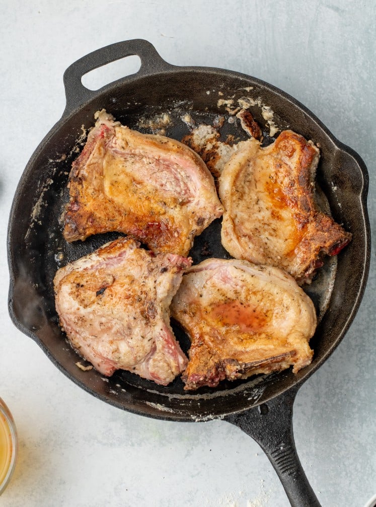 Golden-brown seared pork chops cooking in a cast-iron skillet, showing crispy edges and caramelized surfaces after being browned on both sides.