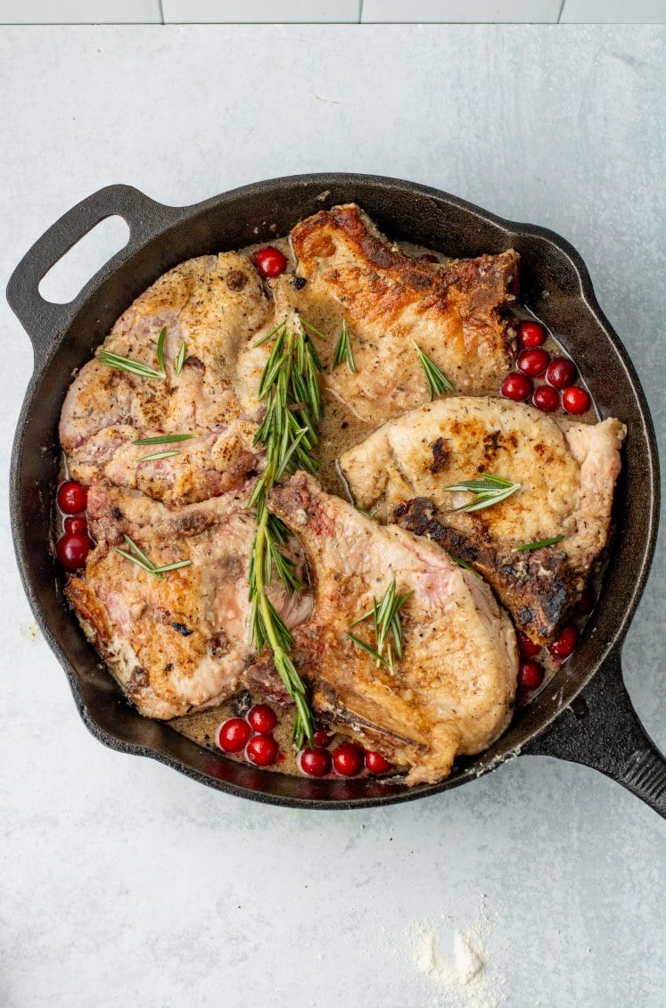 Seared pork chops simmering in a cast-iron skillet with fresh cranberries and rosemary sprigs, finishing in a savory maple cranberry sauce.