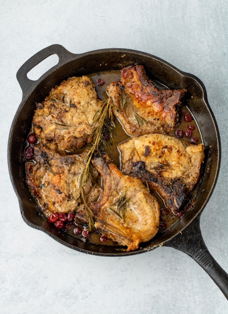 Juicy maple cranberry pork chops cooked to golden perfection in a cast-iron skillet, surrounded by caramelized cranberries and fresh rosemary sprigs.