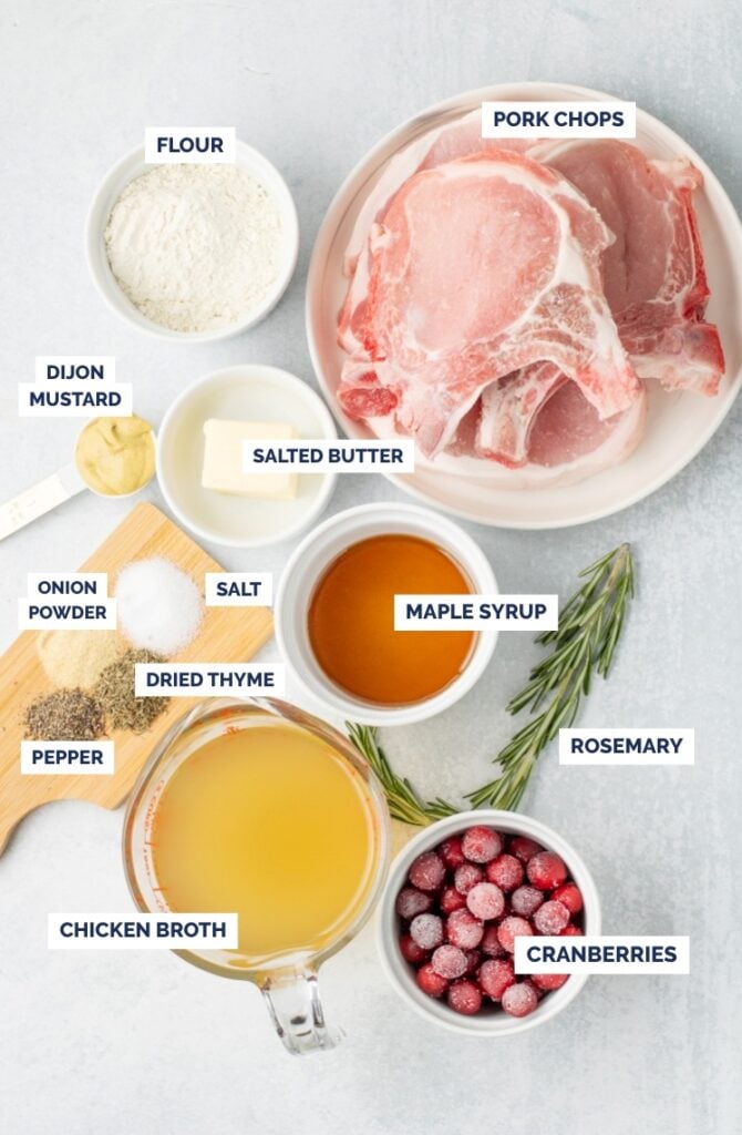 Labeled overhead shot of ingredients for maple cranberry pork chops, including pork chops, cranberries, rosemary, maple syrup, chicken broth, flour, butter, Dijon mustard, dried thyme, onion powder, salt, and pepper.
