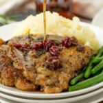 Maple cranberry pork chops served with mashed potatoes and green beans, topped with rich maple sauce being poured from a gravy boat. Text overlay reads ‘Maple Cranberry Pork Chops – Real Balanced.