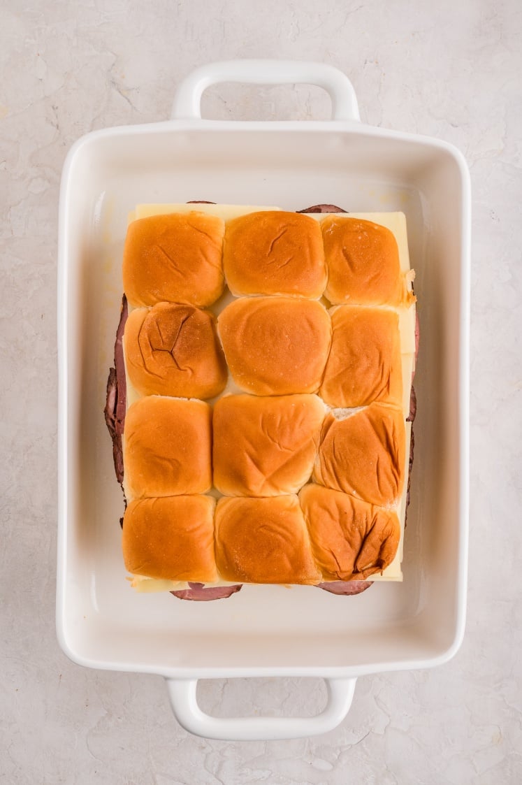 White baking dish with a layer of soft, golden-brown slider buns placed on top of the cheese and roast beef.