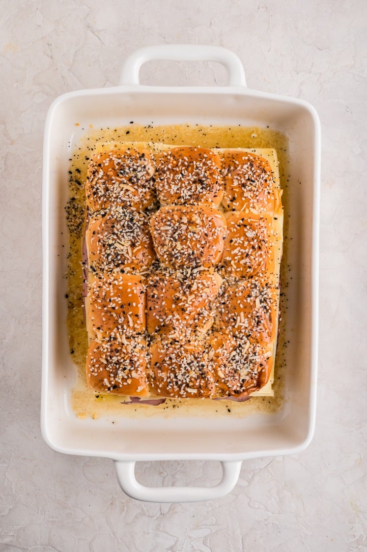 white baking dish filled with slider buns brushed with melted butter and sprinkled with seasoning, sitting in a pool of butter around the edges.