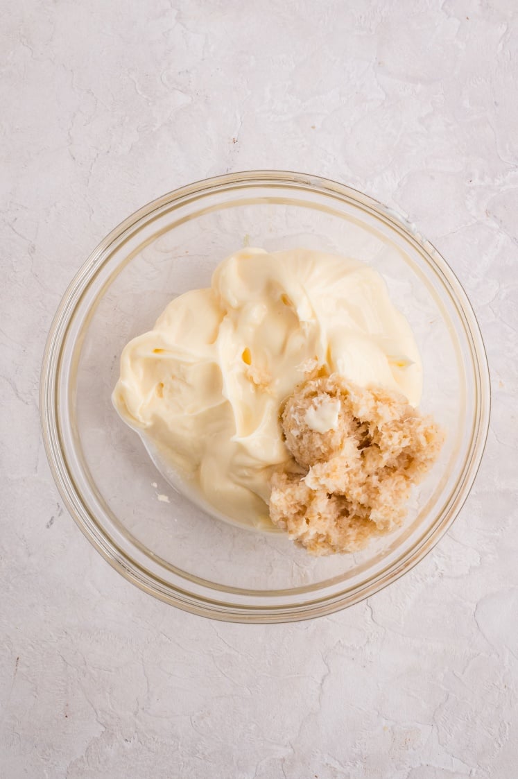 Top view of a glass bowl filled with a scoop of creamy white mayonnaise and a portion of light beige grated or blended mixture beside it.