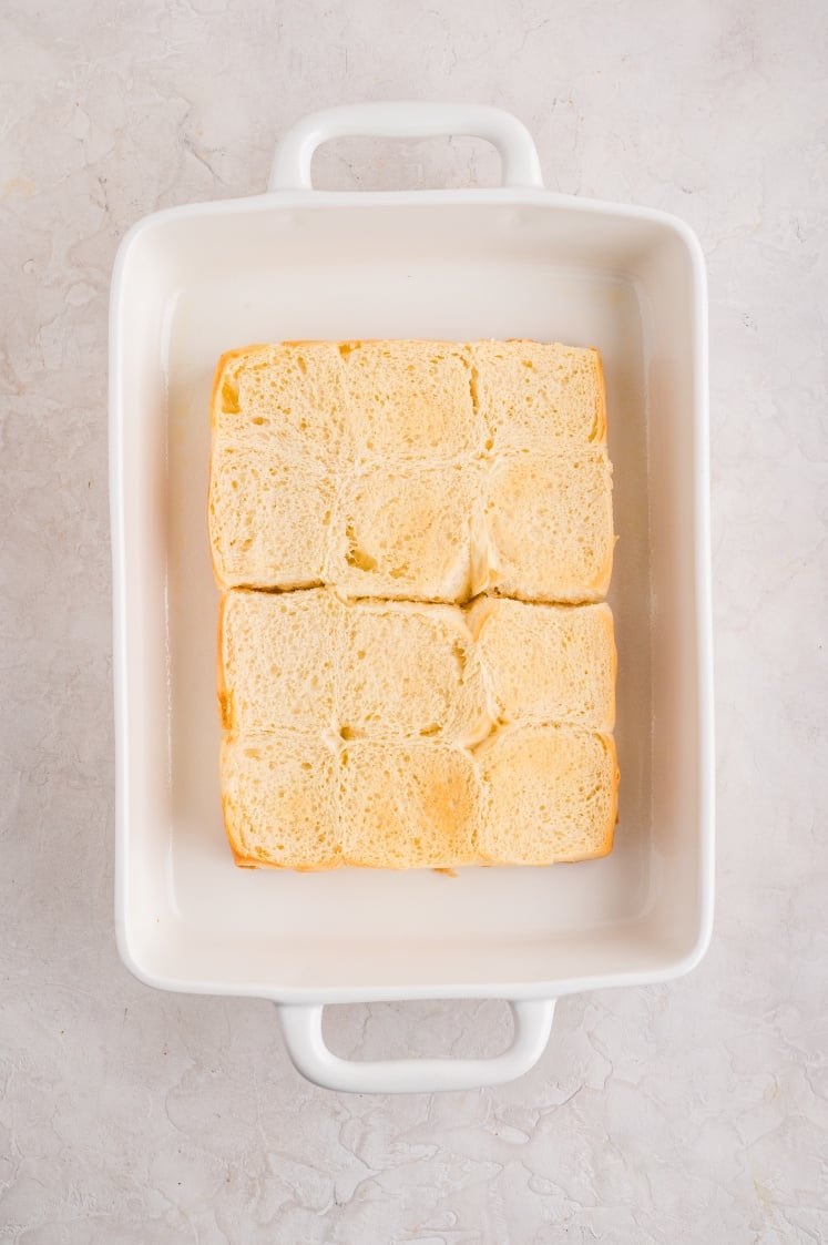 Top view of a white rectangular baking dish with a layer of soft, square bread rolls arranged closely together