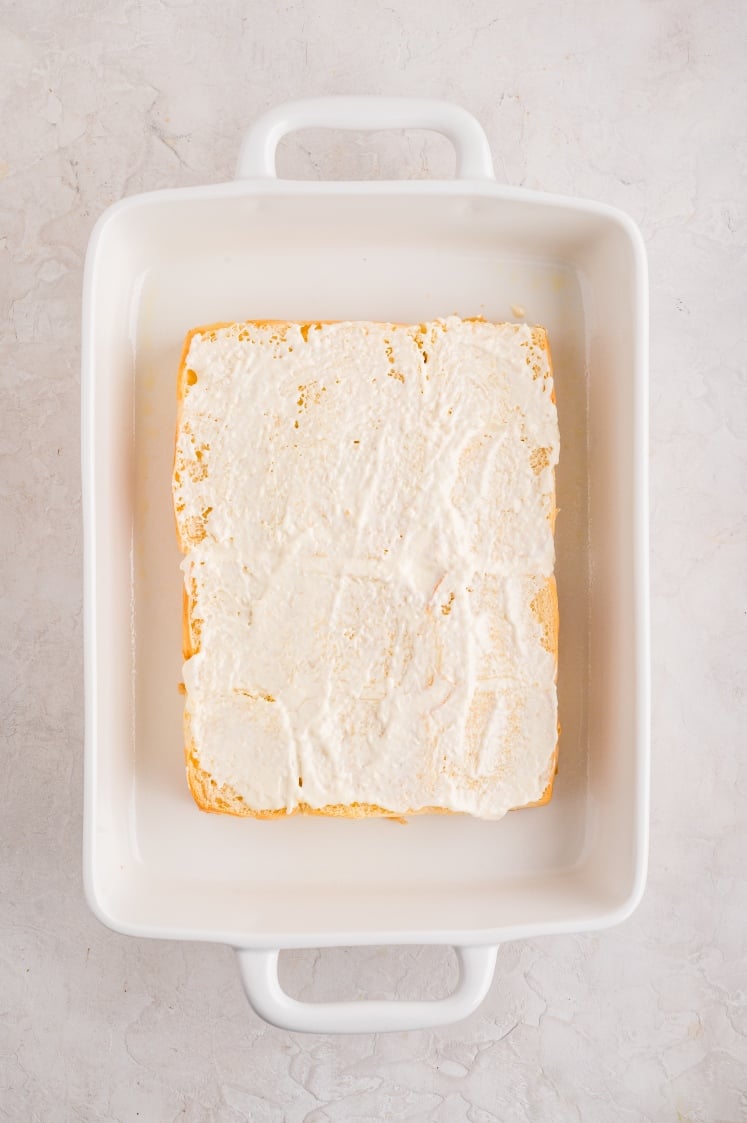 Top view of a white baking dish holding a sheet of bread rolls with a thick, creamy white spread covering the cut surface.