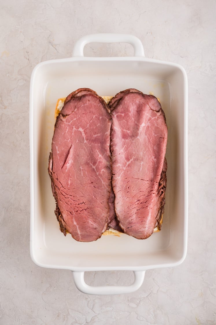 Top view of a white baking dish with two large slices of pink roast beef laid side by side on top of the prepared bread.