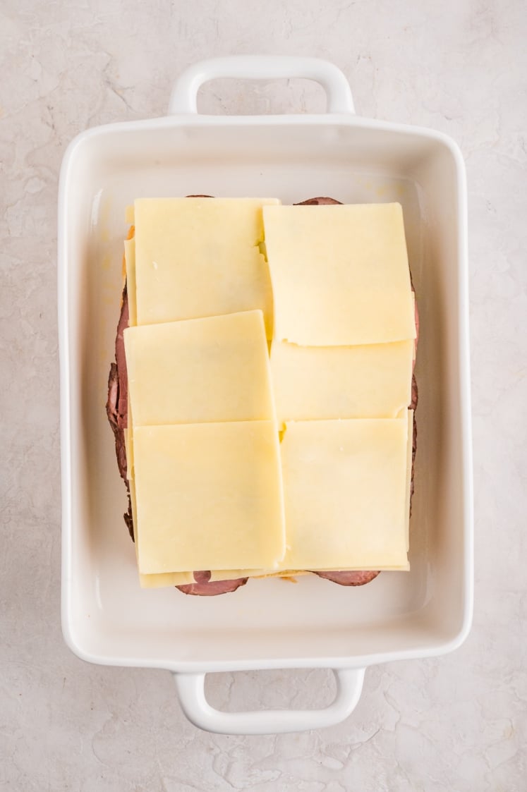 White baking dish with several slices of plain, holeless cheese layered evenly over the Swiss cheese and roast beef.